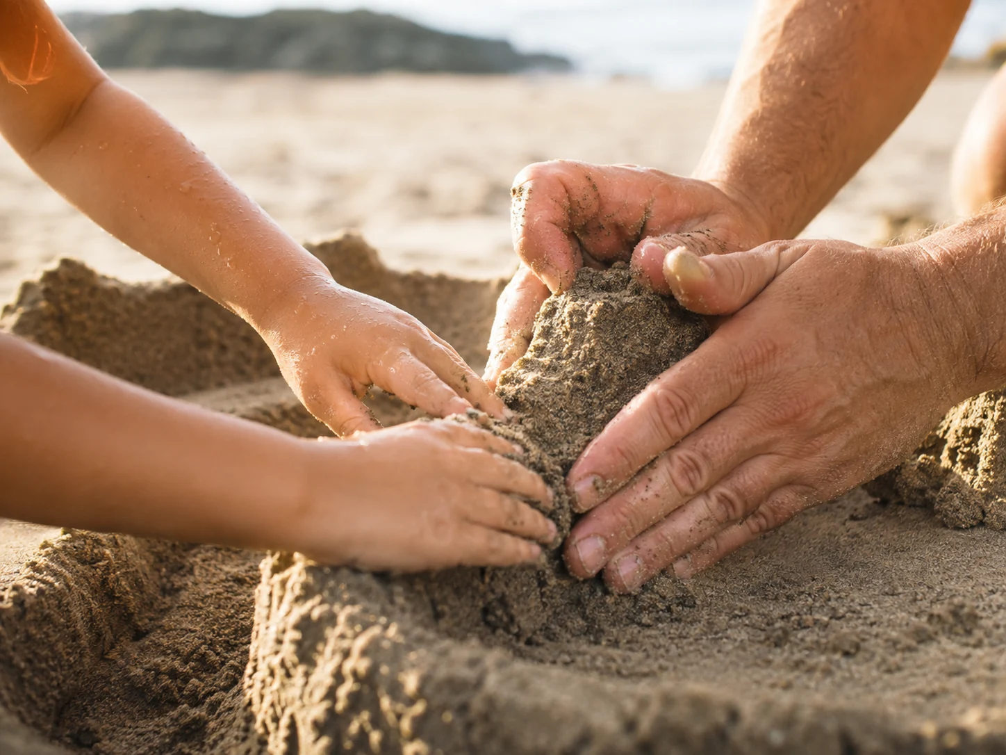 Close up of a child's hands and an adult's hands packing wet sand together on the wall of a sandcastle, the beach and a headland soft behind them.