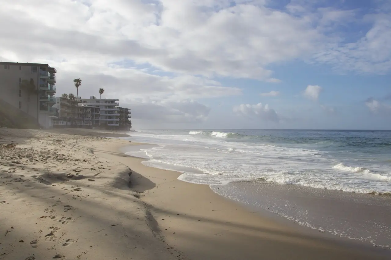 A wide, empty stretch of Laguna sand with foam running up the shore, oceanfront buildings and palms at the far left under a tall bank of broken clouds.