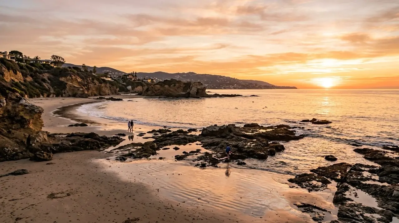 A Laguna cove at sunset, wet sand and tide pools reflecting the orange sky, homes on the bluff at the left and two people walking the waterline in the distance.