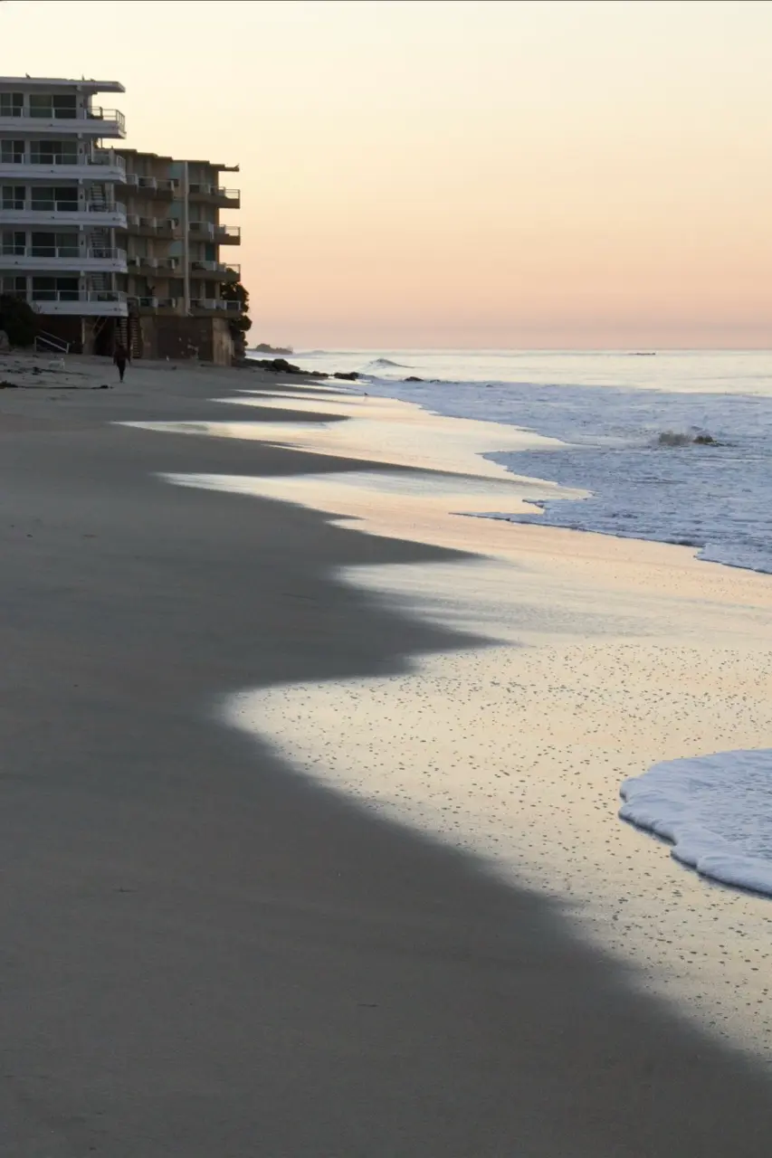 An empty Laguna beach just after dawn, pale pink sky, a scalloped line of foam sliding up the sand, and a white oceanfront building on the bluff at the left.