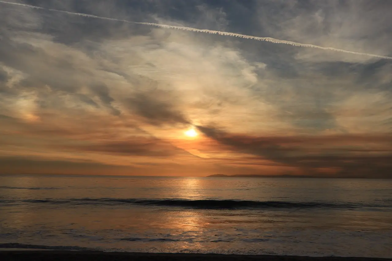 The sun low over the ocean behind long streaked clouds, the water dark and quiet below, with a single contrail crossing the sky.