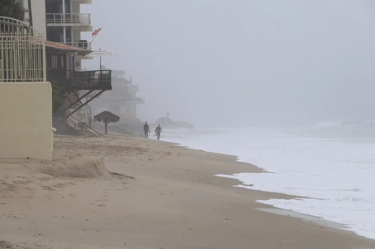 Two distant figures walking a wide beach in heavy coastal fog, a beachfront house and flag softened behind them.
