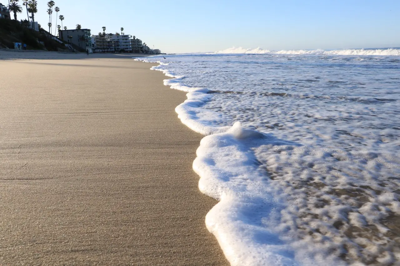 A low view along the Laguna shoreline, sea foam running up firm wet sand toward the camera, palms and hillside homes small in the distance.