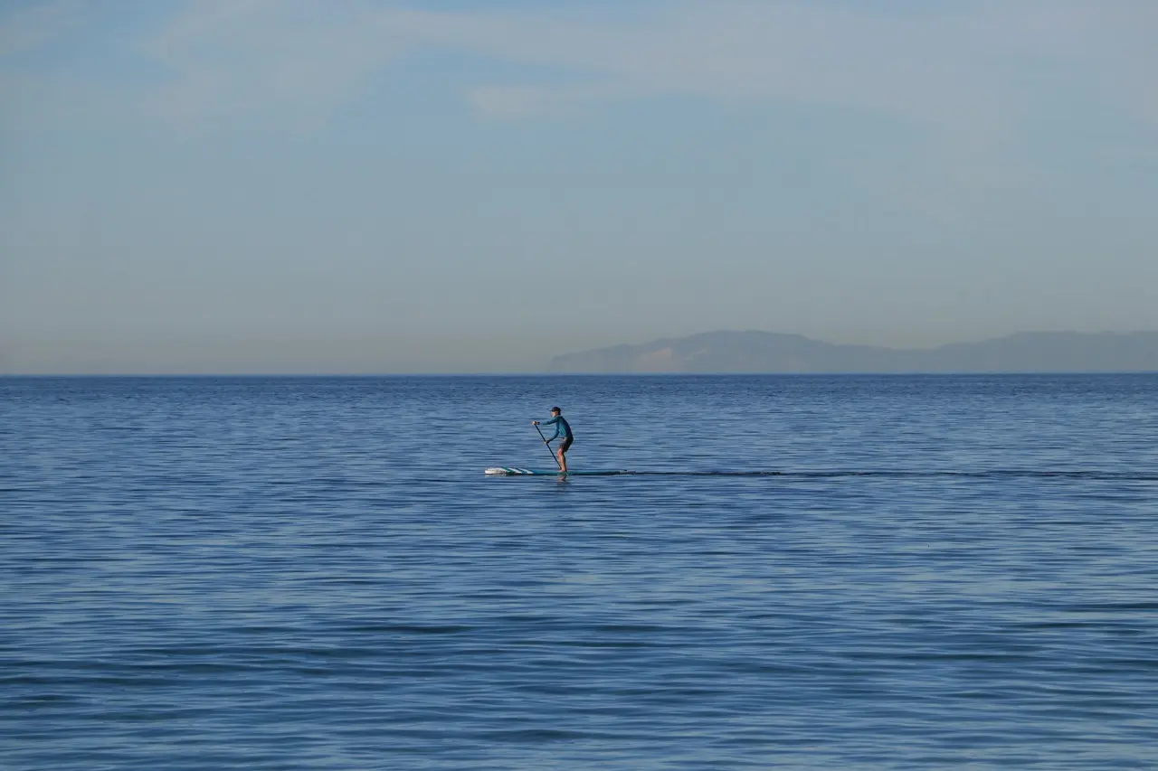 A single stand-up paddleboarder crossing a wide, glassy blue sea, with a hazy island low on the horizon.