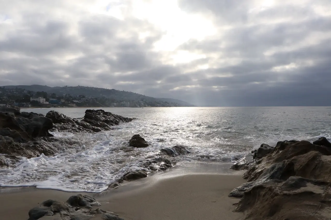 Sunlight breaking through heavy cloud over a silver sea, dark wet rocks in the foreground and the Laguna hillside faint across the water.