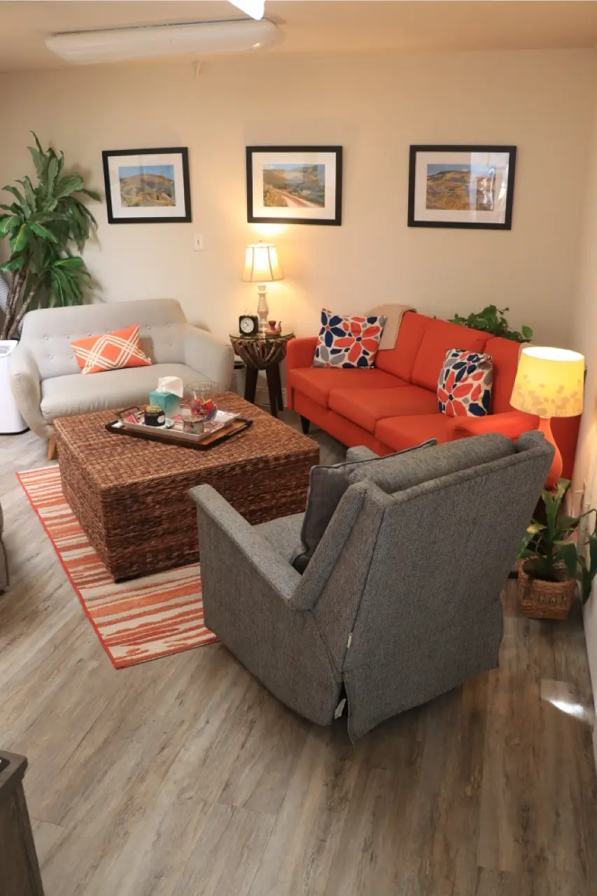 A session room at the practice, an orange sofa and a grey armchair around a woven coffee table, framed landscape photographs on the wall and lamps lit.