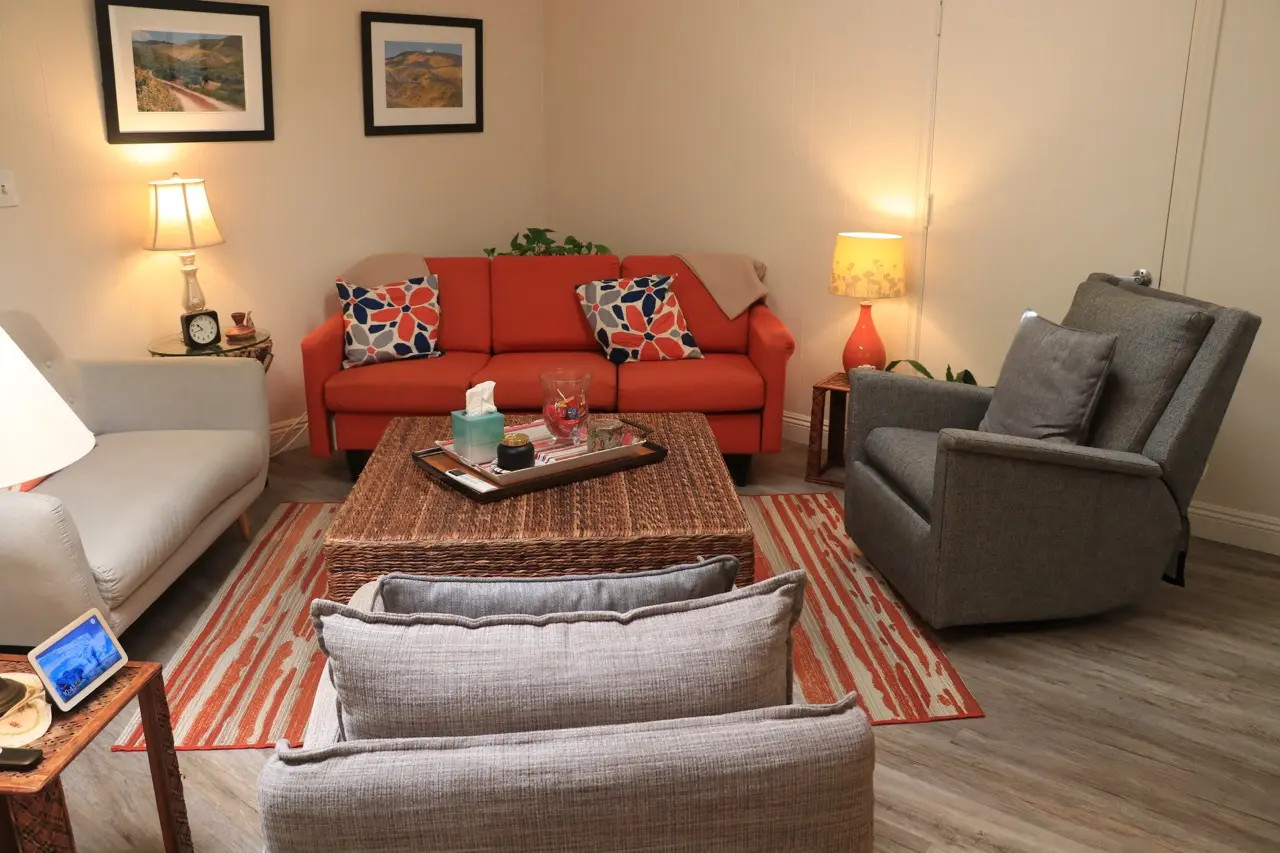 A session room at the practice seen from the doorway, an orange sofa and armchairs around a woven coffee table, table lamps lit and framed landscapes on the wall.