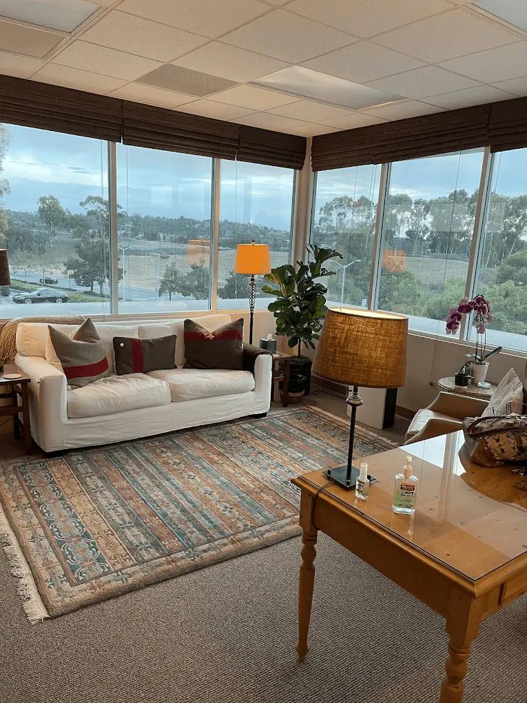 The sister office in Irvine, a corner room with a white sofa, a patterned rug and floor lamps, its windows looking out over trees and low hills.