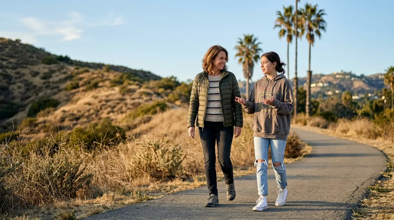 A parent and a teenager walking side by side along a paved trail through golden coastal hills, mid-conversation.