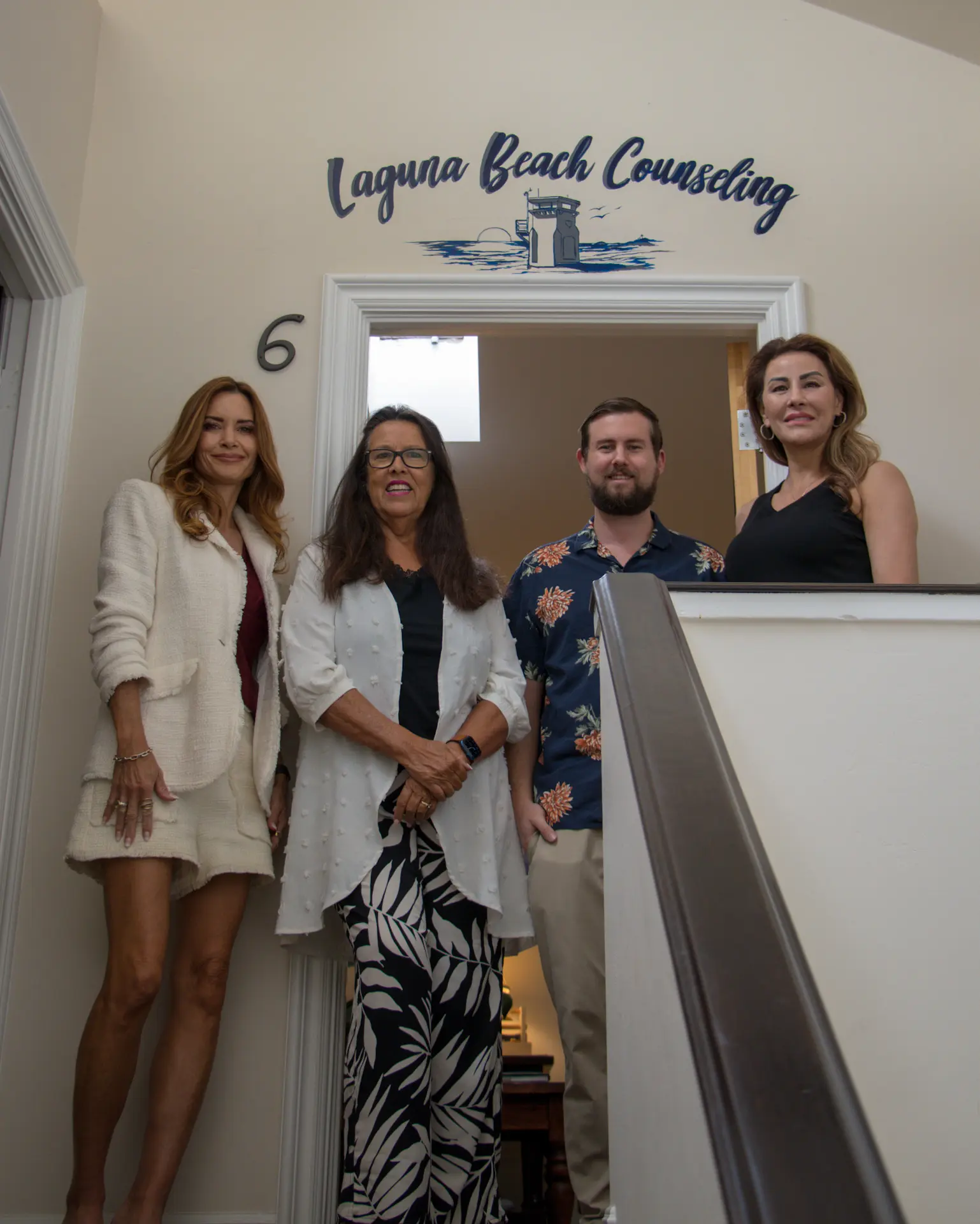 The staircase up to the practice, with the hand-painted Laguna Beach Counseling name and a suite number six on the wall at the top, four of the team standing on the stairs.