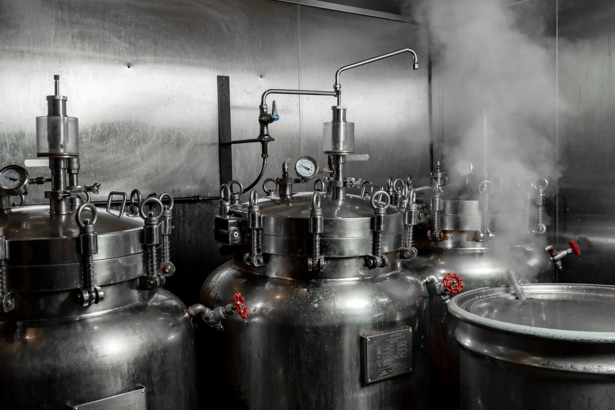 The Tajima commissary kitchen, a row of stainless steel broth vessels with steam rising and red shut-off valves visible